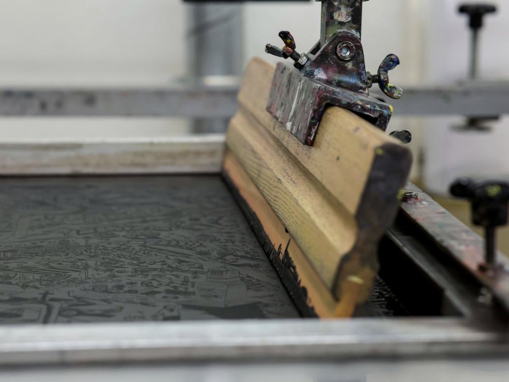 A screen printing press with a squeegee drawing ink across a screen during a production run.