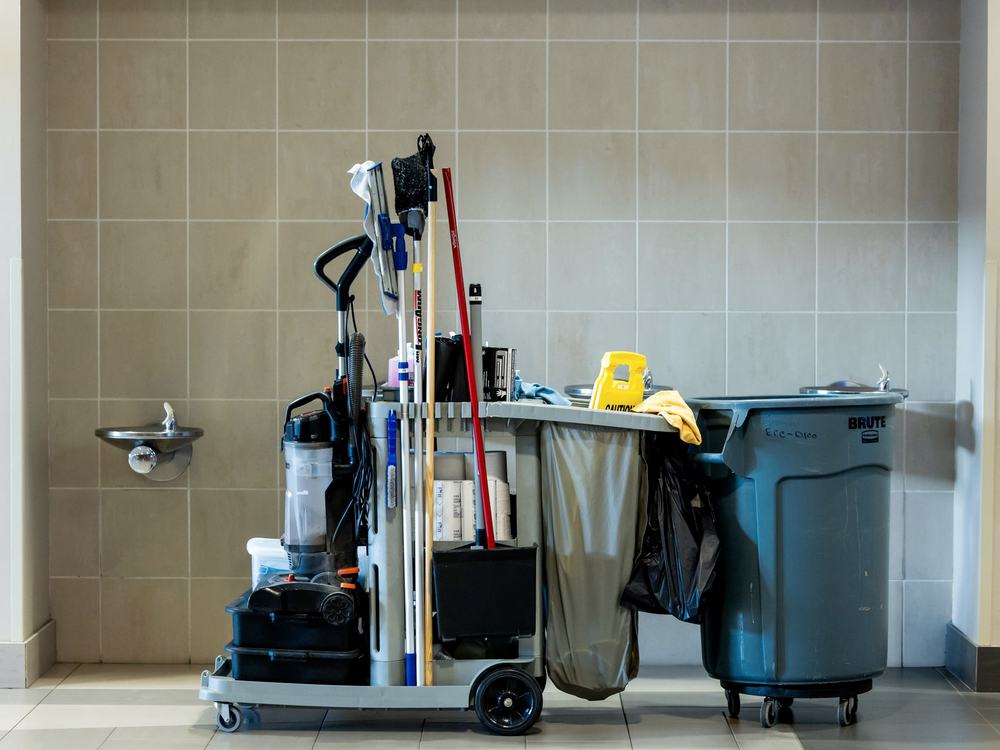 A janitorial cart with cleaning tools, waste bag and a wheeled waste container in a tiled facility corridor.